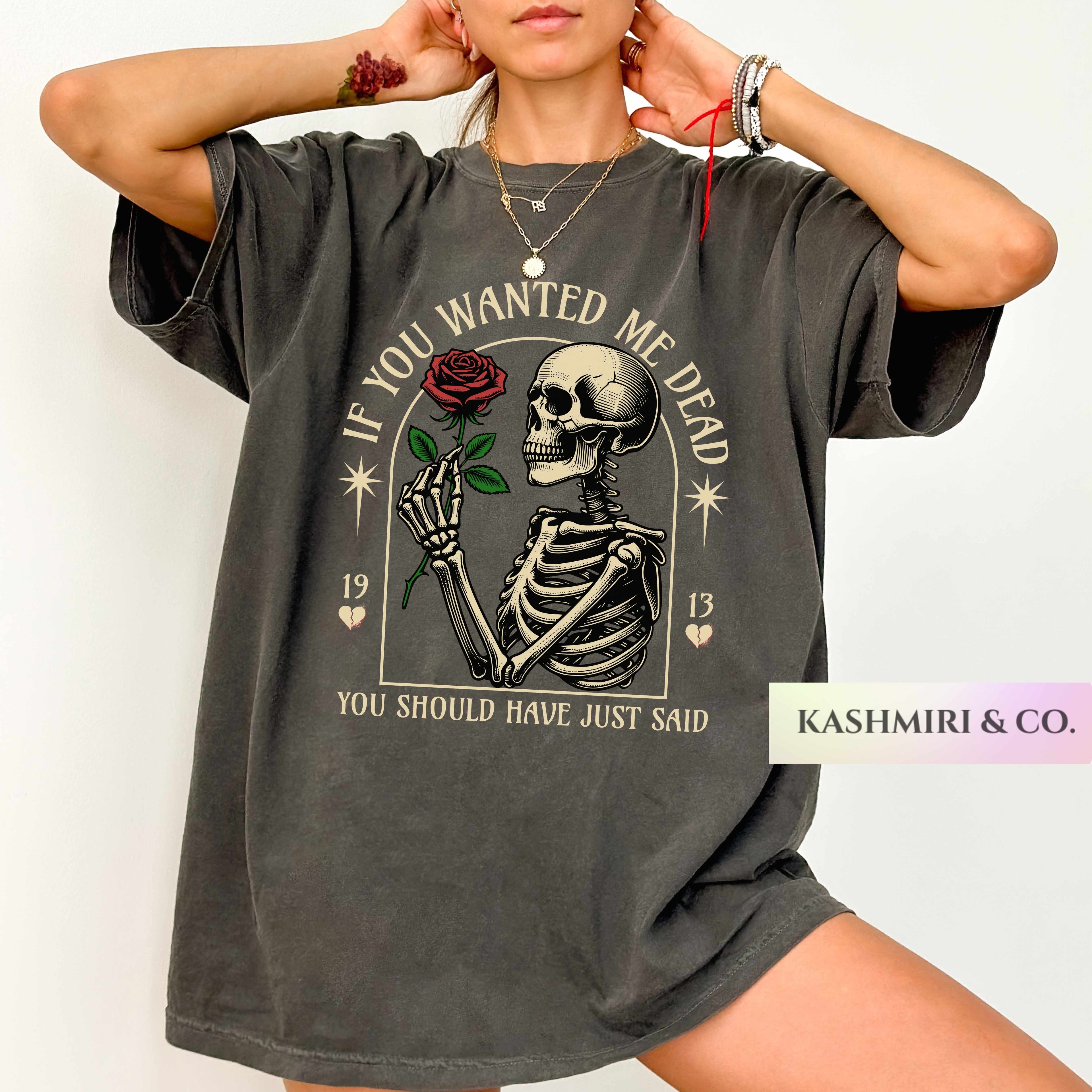 A woman wearing an oversized charcoal gray graphic t-shirt featuring a skeleton holding a red rose with the text 'IF YOU WANTED ME DEAD YOU SHOULD HAVE JUST SAID'. A 'KASHMIRI & CO.