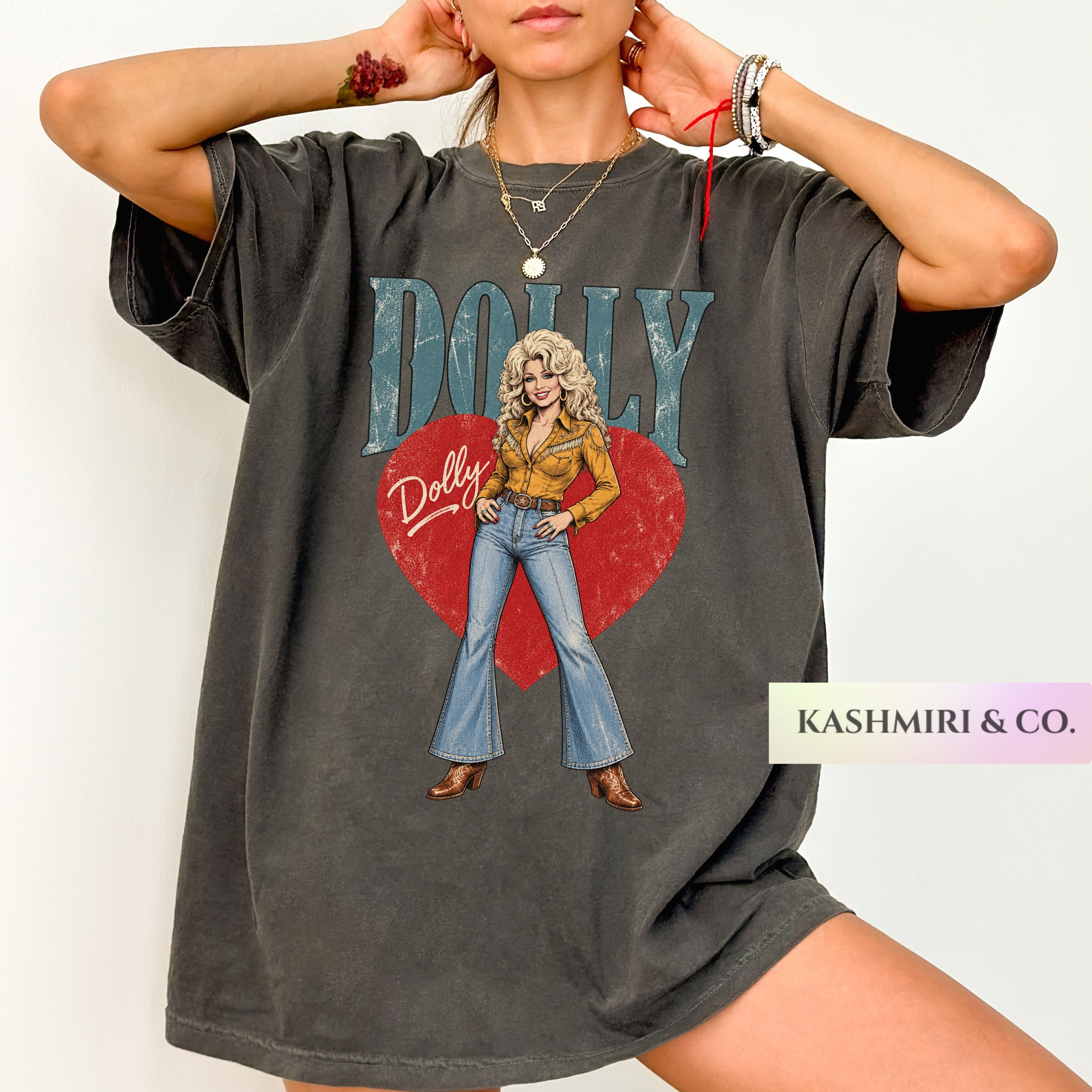 A woman wearing an oversized dark gray graphic T-shirt featuring a vintage-style illustration of Dolly Parton with the text 'DOLLY' and a red heart. The photo is cropped from the neck down against a plain light background, with a 'KASHMIRI & CO.