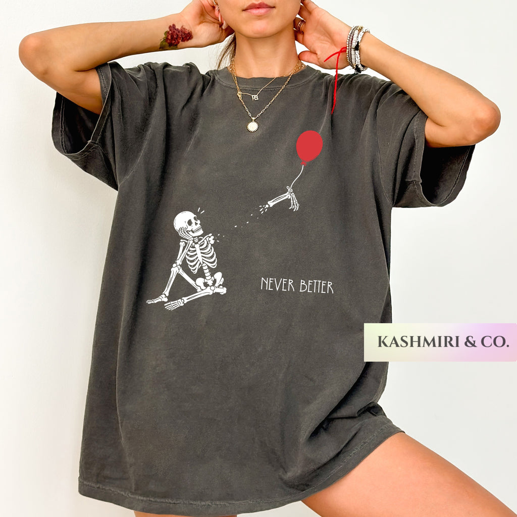 A woman wearing an oversized dark gray graphic t-shirt with a white skeleton pulling a red balloon and the text 'NEVER BETTER'. The brand label 'KASHMIRI & CO.