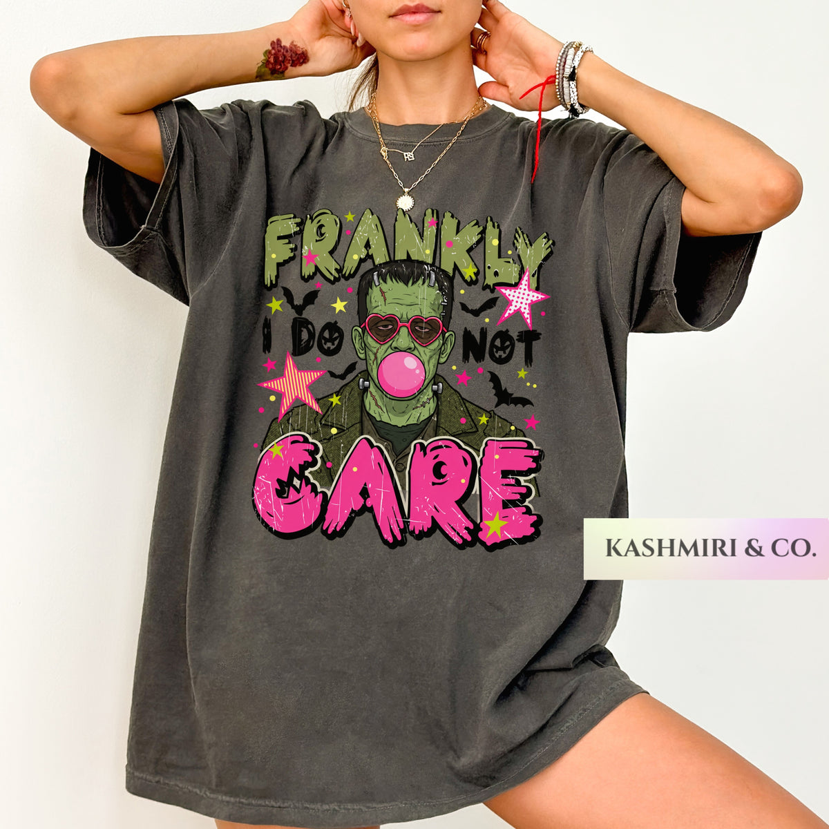 A woman wearing an oversized charcoal gray graphic t-shirt with a Frankenstein-themed print that reads 'FRANKLY I DO NOT CARE' in green and pink lettering. A brand label for 'KASHMIRI & CO.