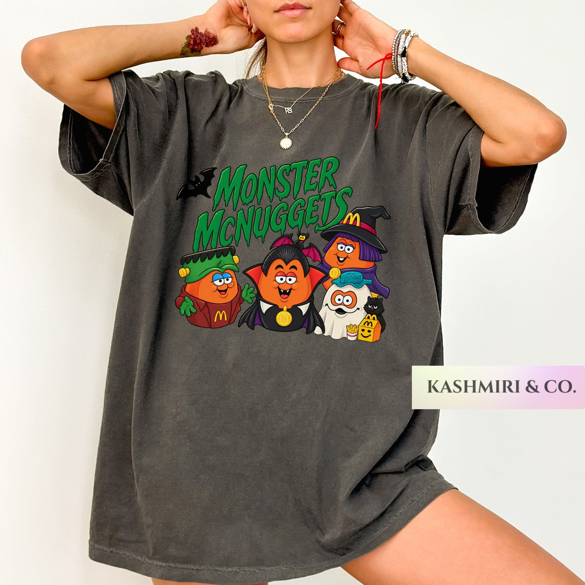 A woman wearing an oversized dark gray t-shirt with a colorful graphic of cartoon characters dressed as Halloween and the text 'Monster McNuggels'. The brand name 'KASHMIRI & CO.