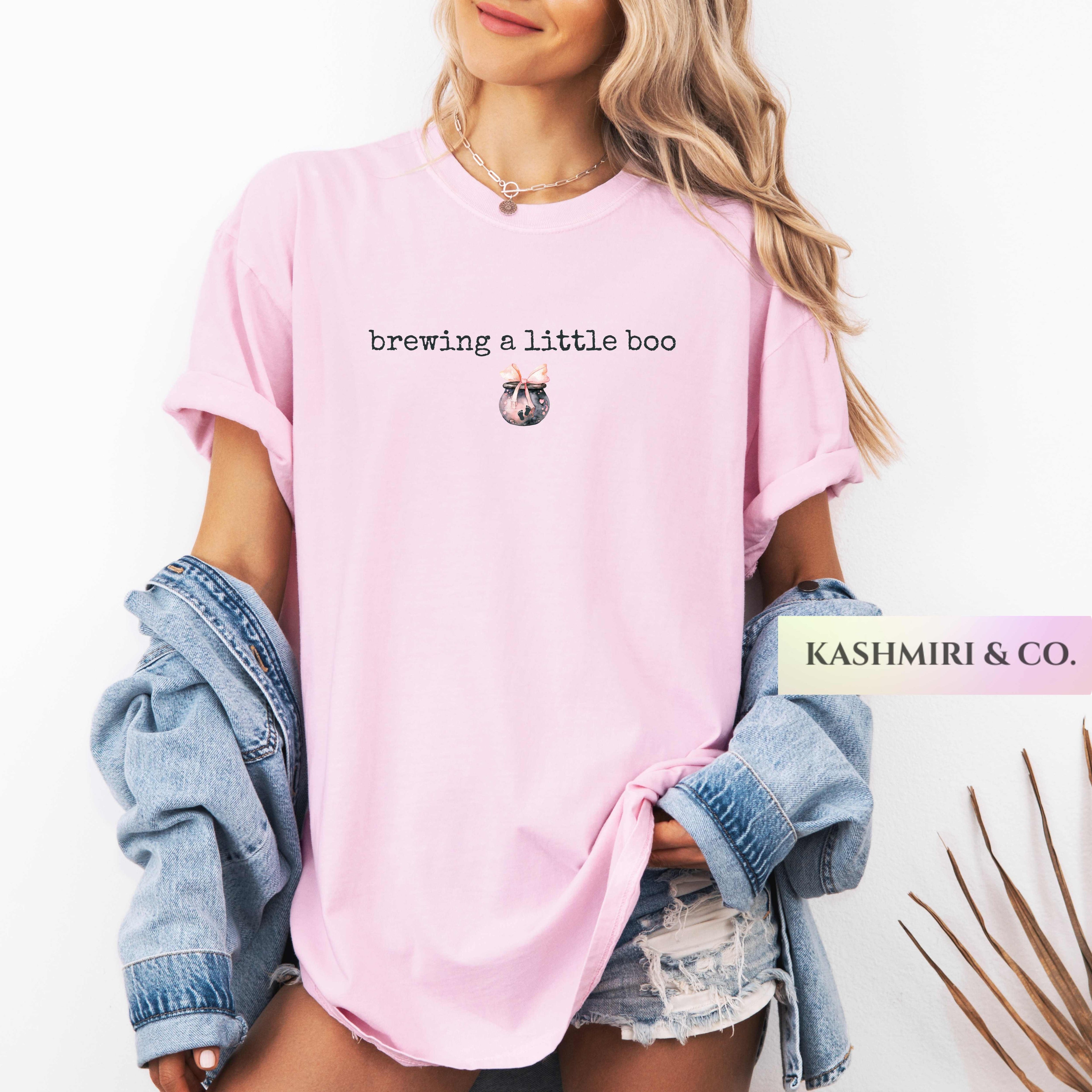 A woman wearing a light pink graphic t-shirt with the text 'brewing a little boo' and a small potion brew illustration. She has a light blue denim jacket draped off her shoulders and is wearing denim shorts.