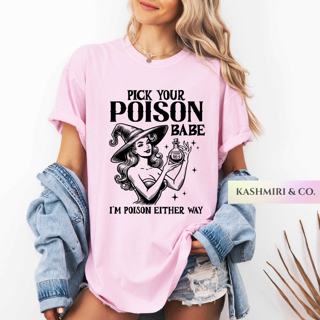 A woman wearing a light pink graphic t-shirt with a black print of a witch and the text 'PICK YOUR POISON BABE I'M POISON EITHER WAY'. She has a light blue denim jacket draped off her shoulders and is wearing denim shorts.