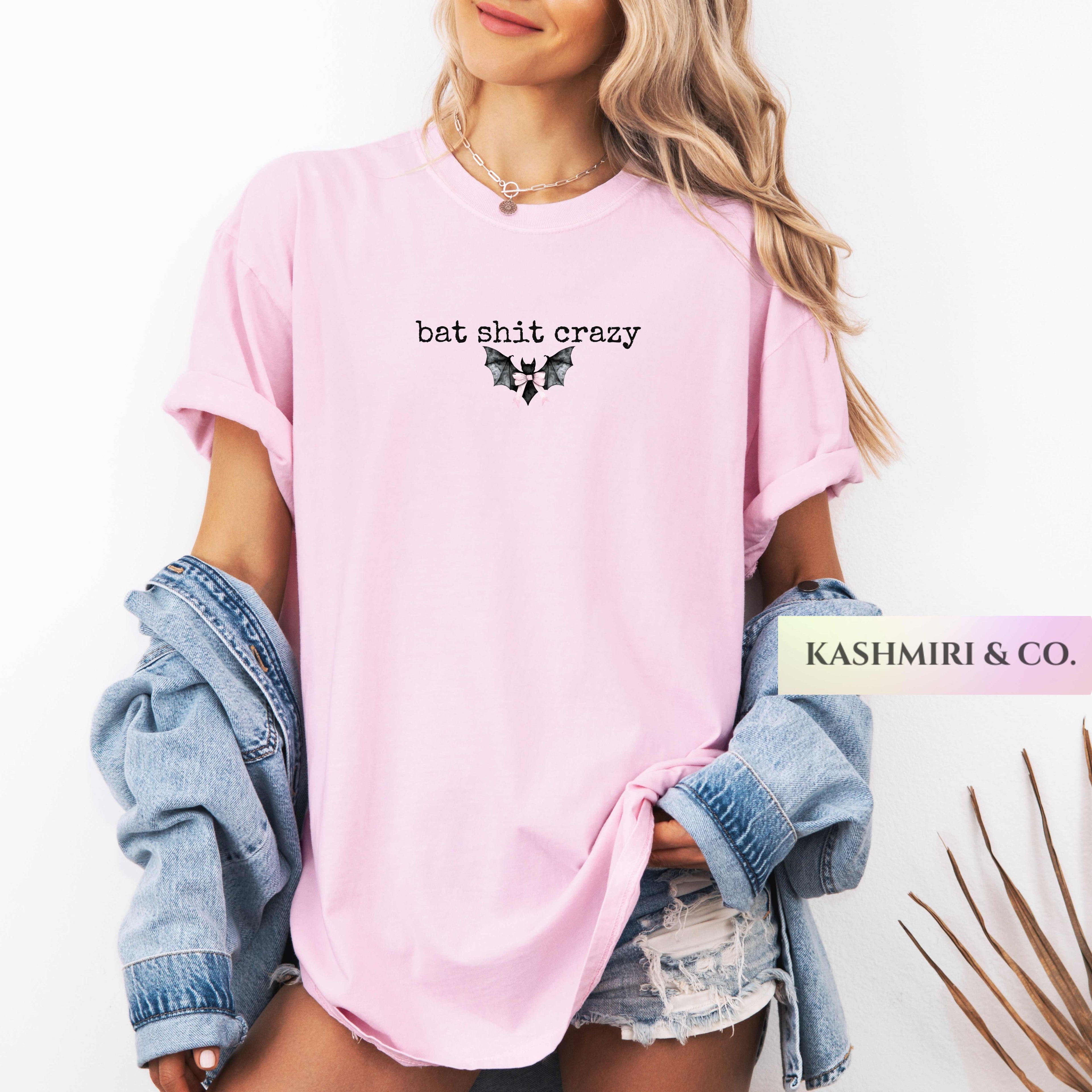 A woman wearing a light pink oversized T-shirt with the text 'bat shit crazy' and a small black graphic on the chest, styled with a light blue denim jacket draped off her shoulders and distressed denim shorts. A brand label reads 'KASHMIRI & CO.