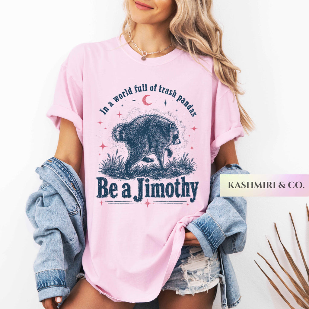 A woman wearing a light pink graphic T-shirt with a bear illustration and the text 'In a world full of trash pandas Be a Jimothy'. She has a light blue denim jacket draped off her shoulders and is wearing distressed denim shorts against a clean white background.