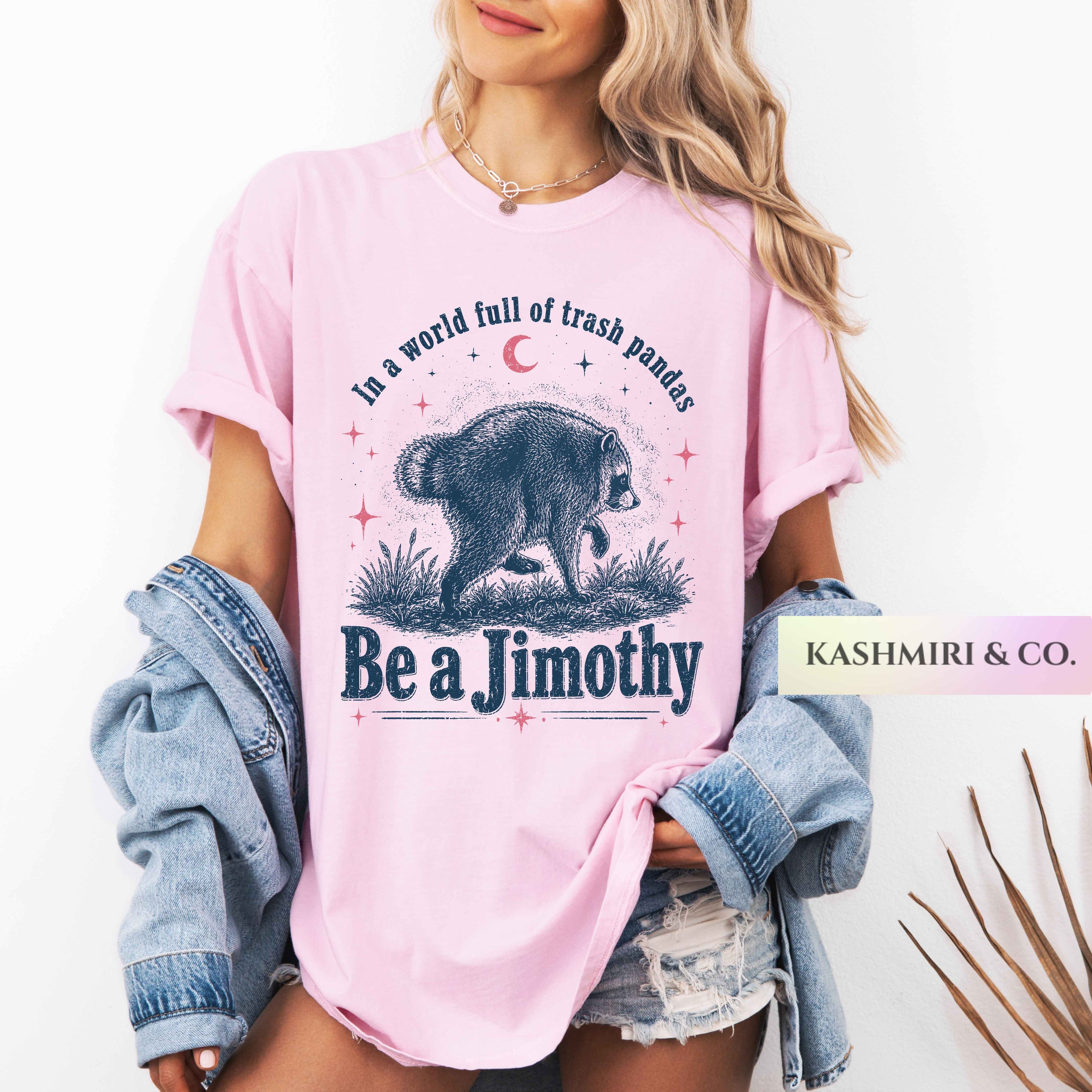 A woman wearing a light pink graphic T-shirt with a bear illustration and the text 'In a world full of trash pandas Be a Jimothy'. She has a light blue denim jacket draped off her shoulders and is wearing distressed denim shorts against a clean white background.
