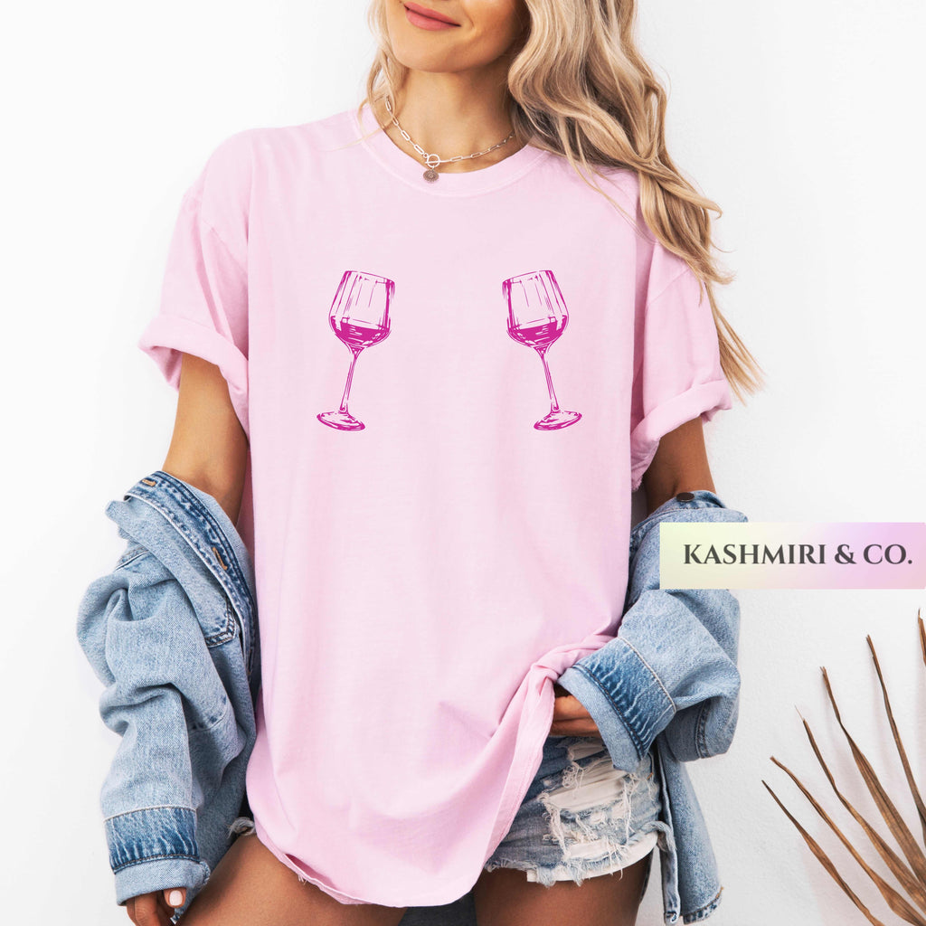 A woman wearing a light pink oversized T-shirt with two pink wine glass graphics on the front, styled with a light blue denim jacket draped off her shoulders and distressed denim shorts. A brand label 'KASHMIRI & CO.