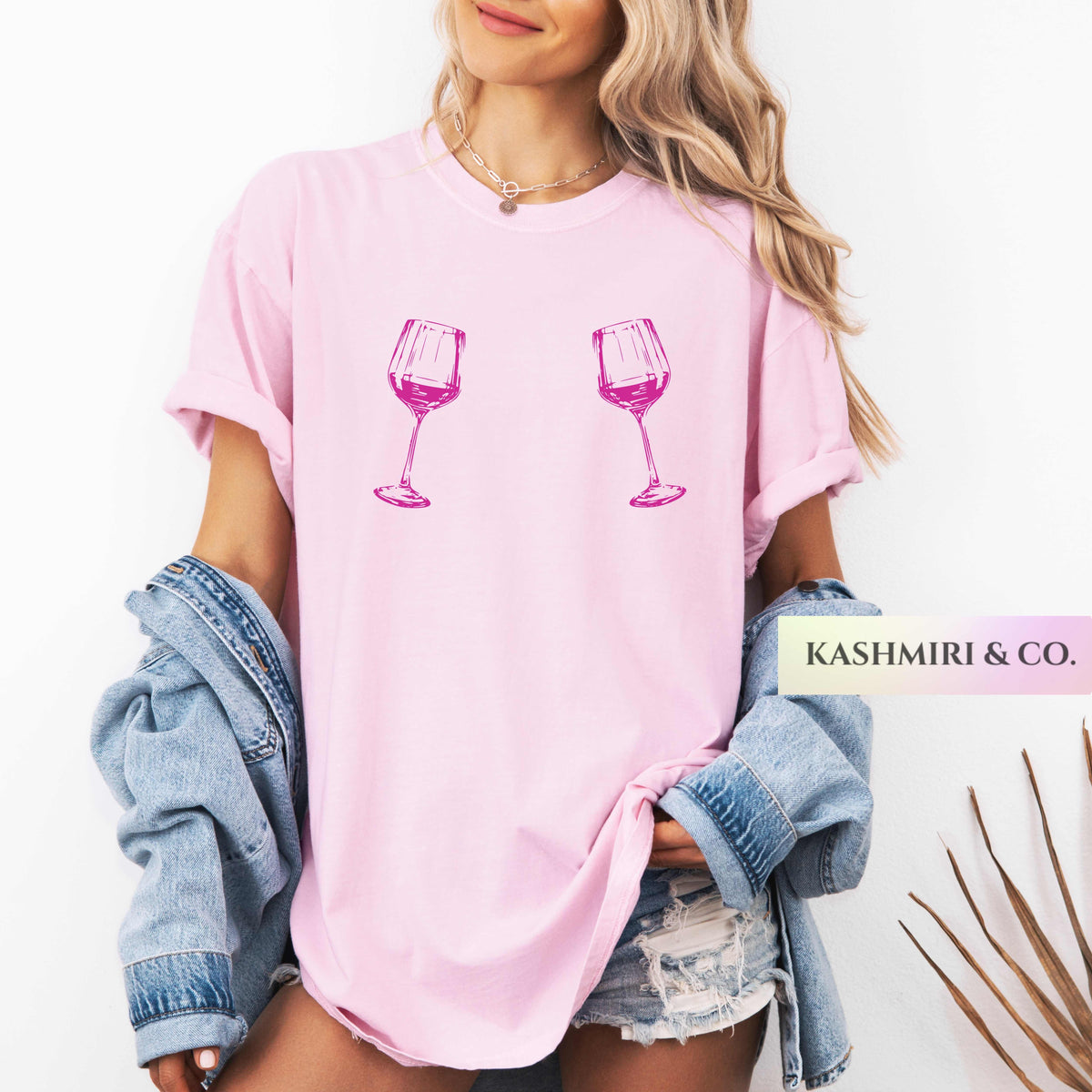 A woman wearing a light pink oversized T-shirt with two pink wine glass graphics on the front, styled with a light blue denim jacket draped off her shoulders and distressed denim shorts. A brand label 'KASHMIRI & CO.