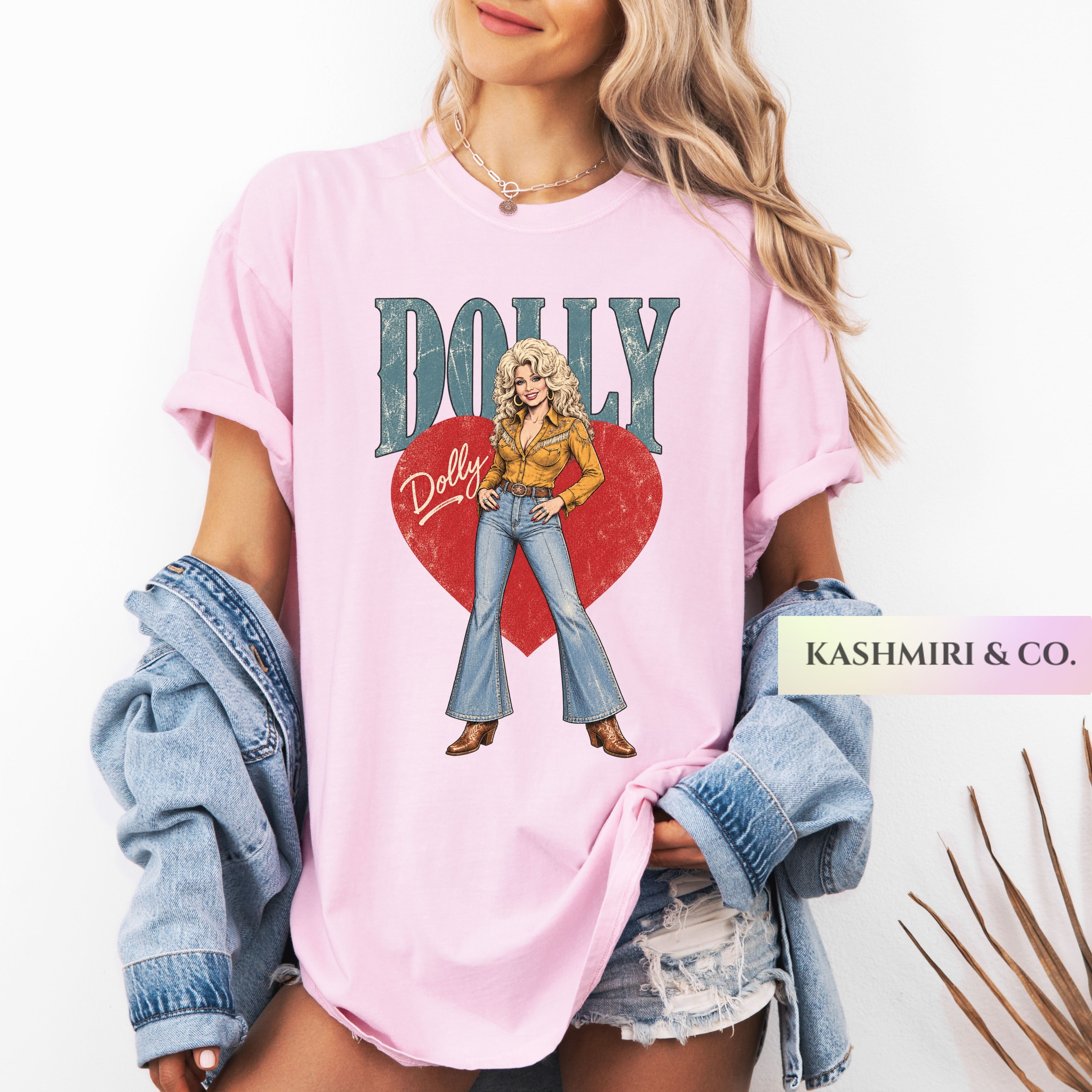 A woman wearing a light pink graphic T-shirt with a retro-style Dolly Parton illustration and text, styled with a denim jacket draped off the shoulders and distressed denim shorts. A brand label reading 'KASHMIRI & CO.