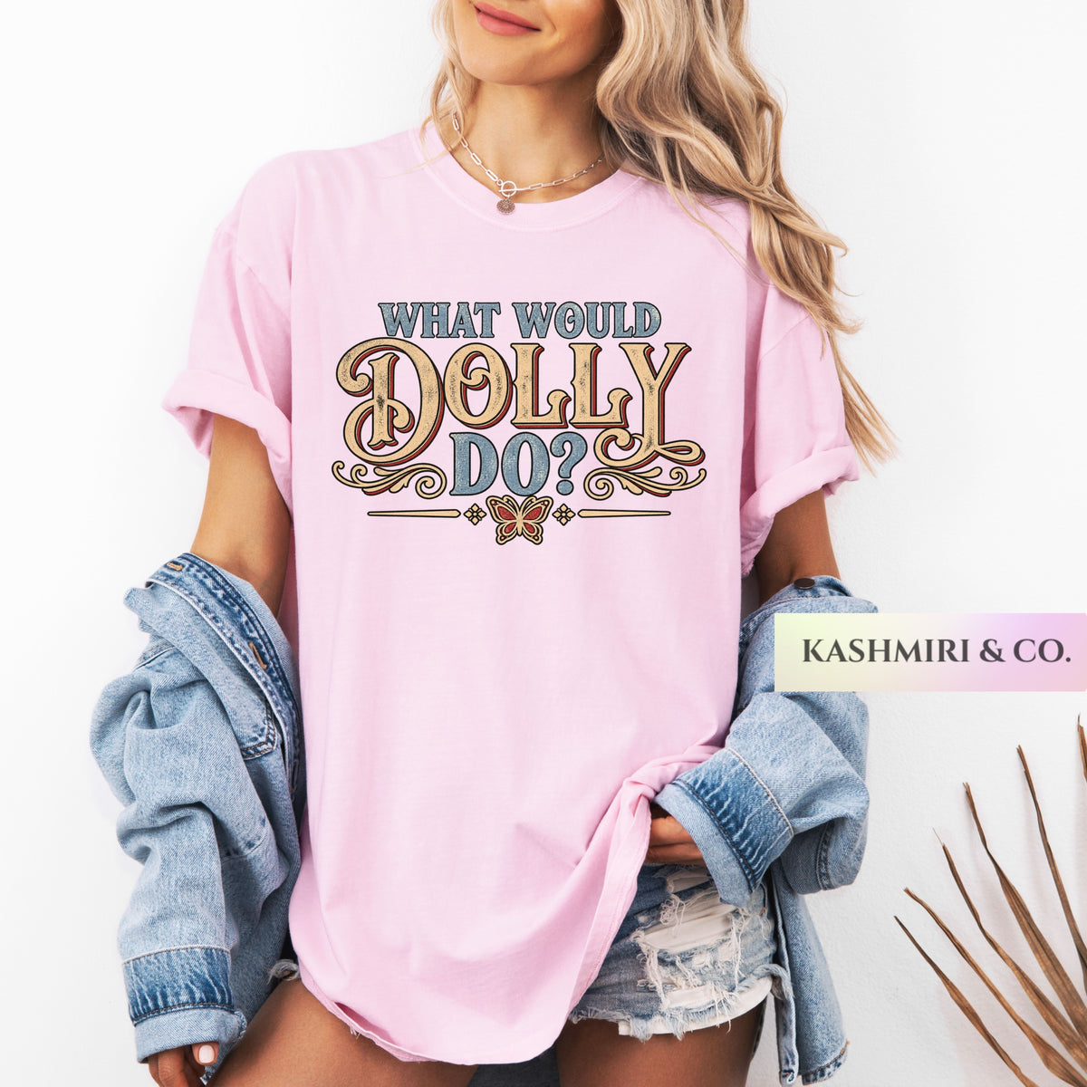 A woman wearing a light pink graphic T-shirt with the text 'WHAT WOULD DOLLY DO?' in decorative lettering. She has a light blue denim jacket draped off her shoulders and is wearing distressed denim shorts against a plain white background.