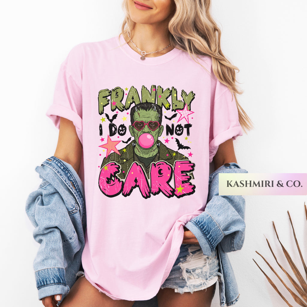 A woman wearing a light pink graphic t-shirt with a Frankenstein-themed design that reads 'FRANKLY I DO NOT CARE' in green, black, and hot pink lettering. She has a light blue denim jacket draped off her shoulders and is wearing distressed denim shorts.