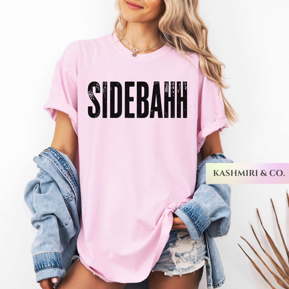 A woman wearing a light pink oversized T-shirt with the word 'SIDEBAHH' printed in distressed black letters across the chest. She has a light blue denim jacket draped off her shoulders and is wearing distressed denim shorts against a clean white background.