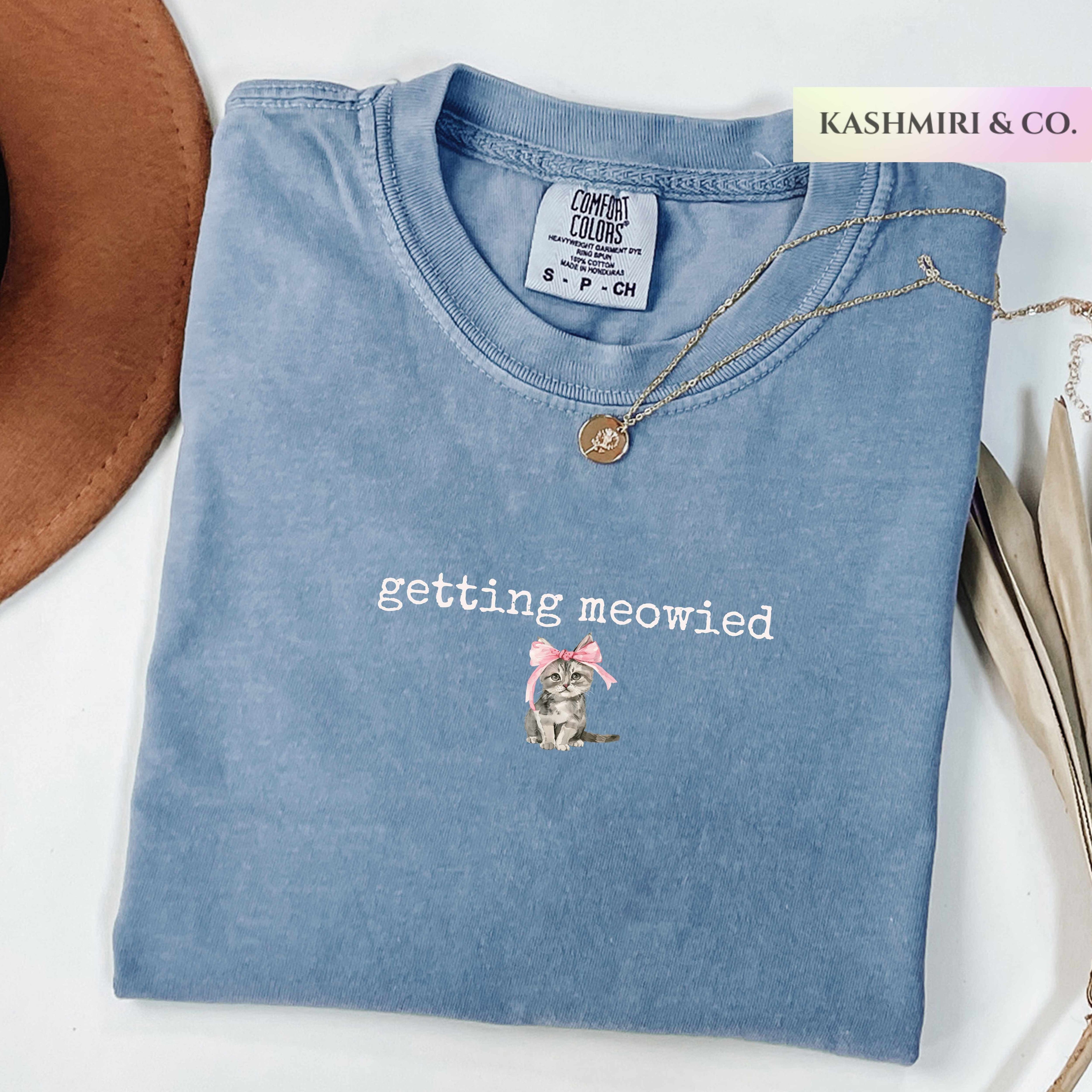 Folded blue t-shirt with 'getting meowied' text and cat illustration, brand 'Kashmiri & Co.' visible.