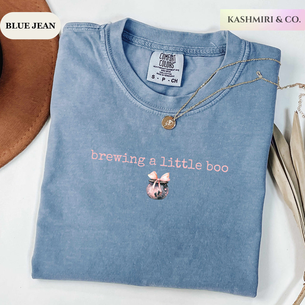 A folded light blue t-shirt with a mineral wash, featuring the text 'brewing a little boo' in pink and a small potion brew graphic. The shirt is styled with a gold necklace and a brown hat on a white background, with text overlays reading 'BLUE JEAN' and 'KASHMIRI & CO.