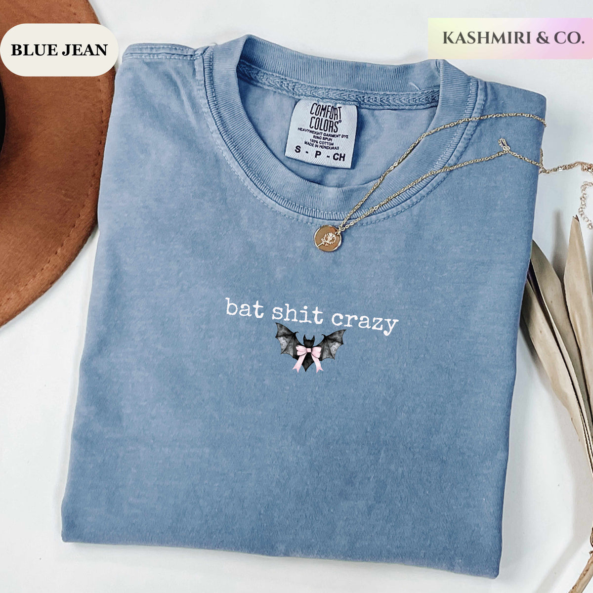 A folded light blue t-shirt with a mineral wash finish, featuring the text 'bat shit crazy' and a small bat graphic on the front. The shirt is styled with a gold necklace and a brown hat on a white surface, with text overlays reading 'BLUE JEAN' and 'KASHMIRI & CO.