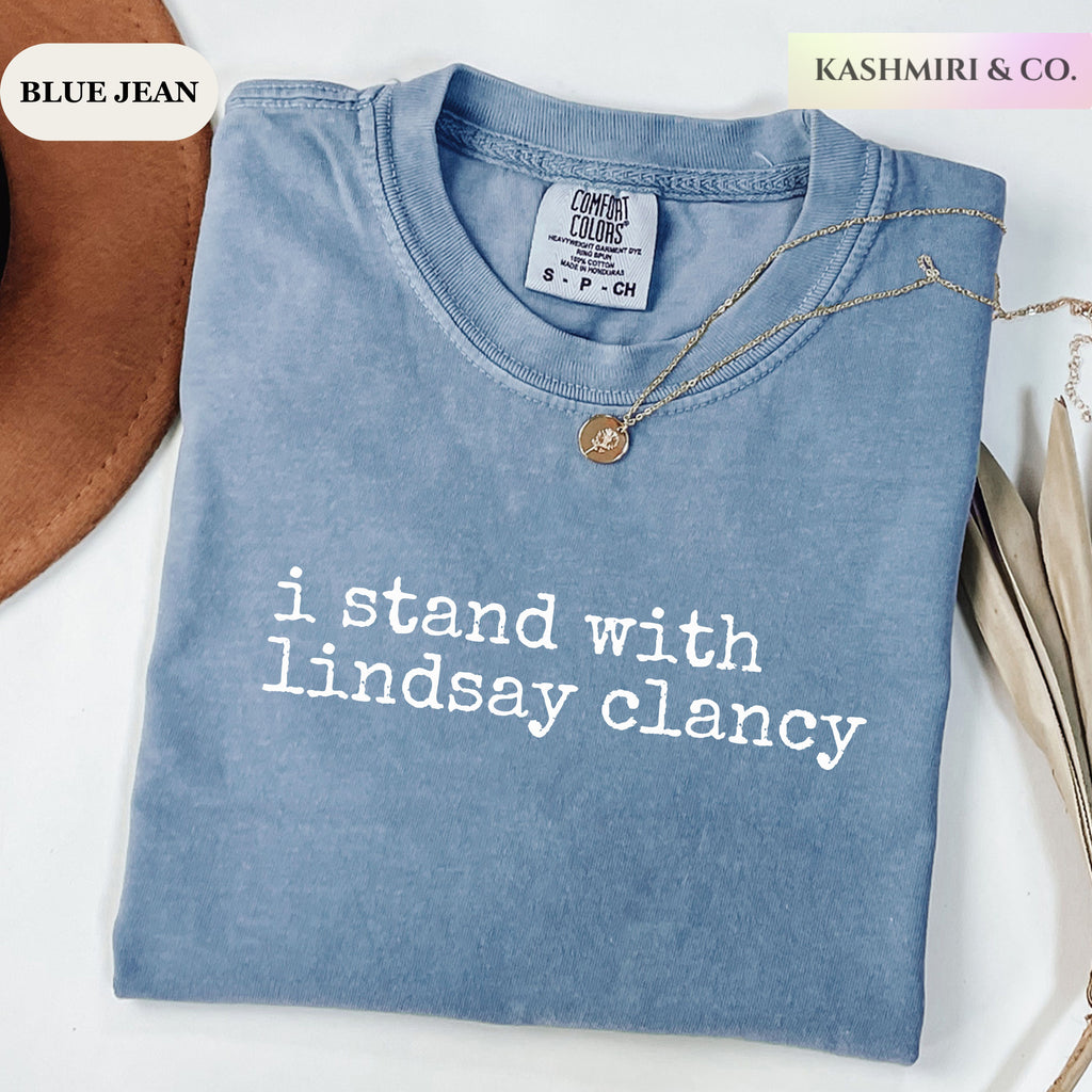 A folded light blue denim T-shirt with white text reading 'i stand with lindsay clancy' on the front, styled on a white surface with a gold necklace and partial props. Brand labels read 'BLUE JEAN' and 'KASHMIRI & CO.