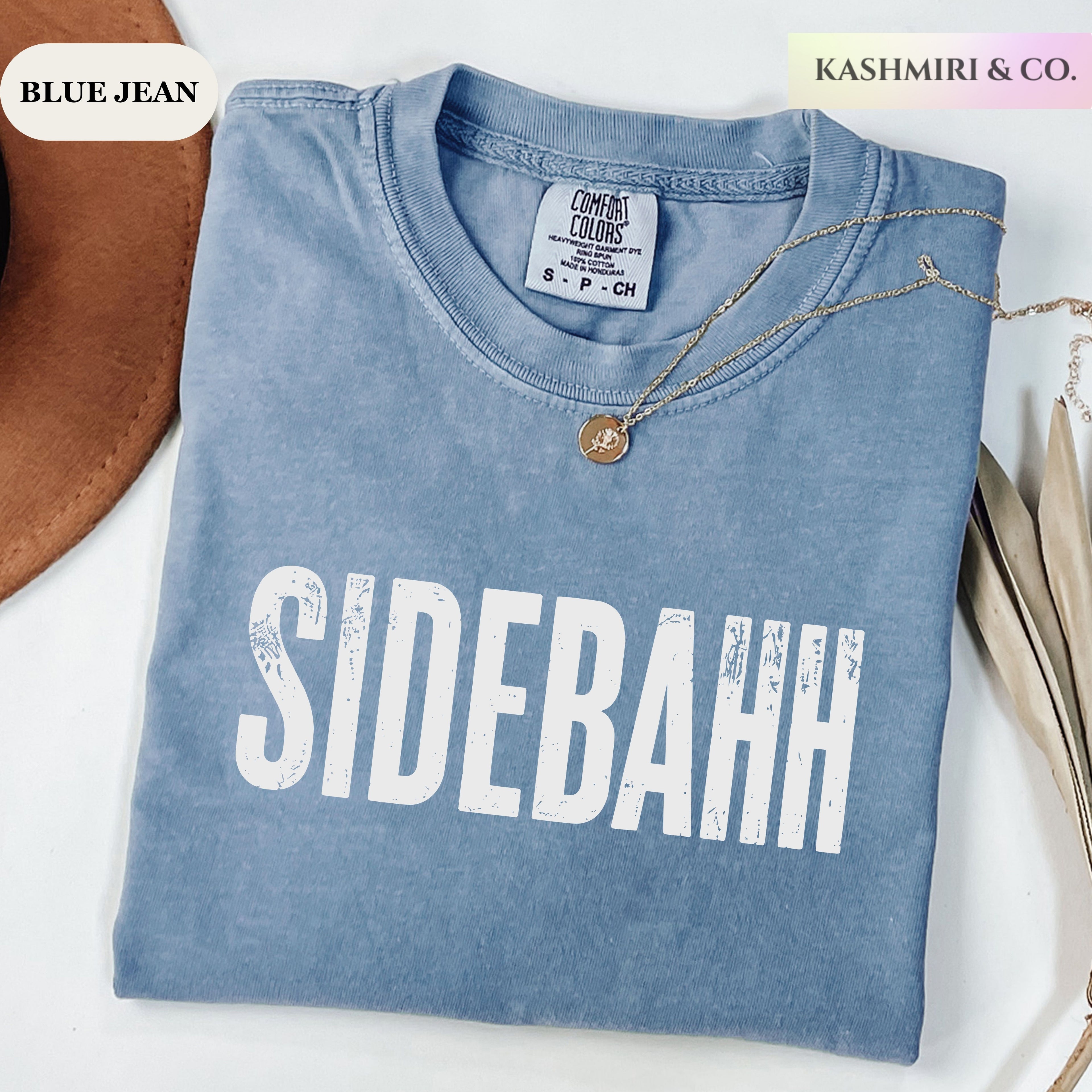A folded light blue denim T-shirt with the word 'SIDEBAHH' printed in large distressed white letters across the front. The shirt is styled on a white surface with a gold necklace draped over the collar, a brown hat edge on the left, and decorative leaves on the right.