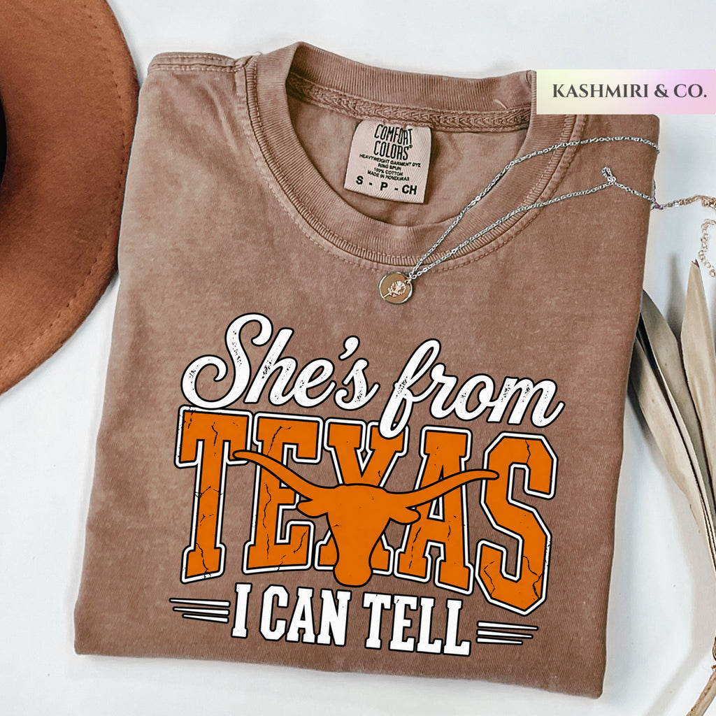A folded brown t-shirt with a graphic that reads 'She's from TEXAS I CAN TELL' in white and orange, styled on a white surface with a brown hat, a silver necklace, and a 'KASHMIRI & CO.' tag.