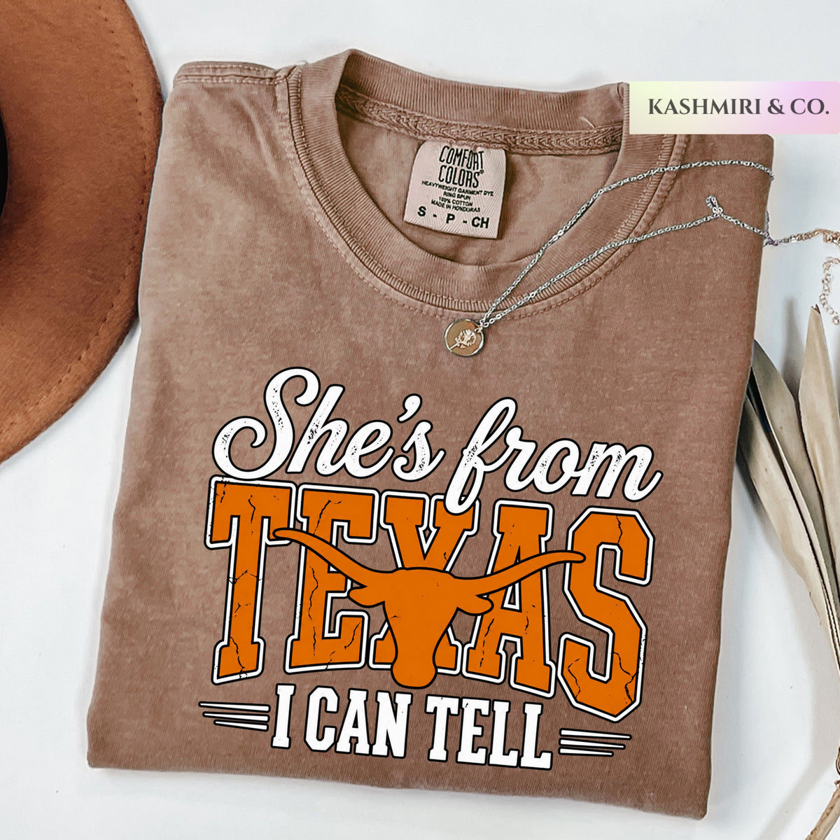 A folded brown t-shirt with a graphic that reads 'She's from TEXAS I CAN TELL' in white and orange, styled on a white surface with a brown hat, a silver necklace, and a 'KASHMIRI & CO.' tag.