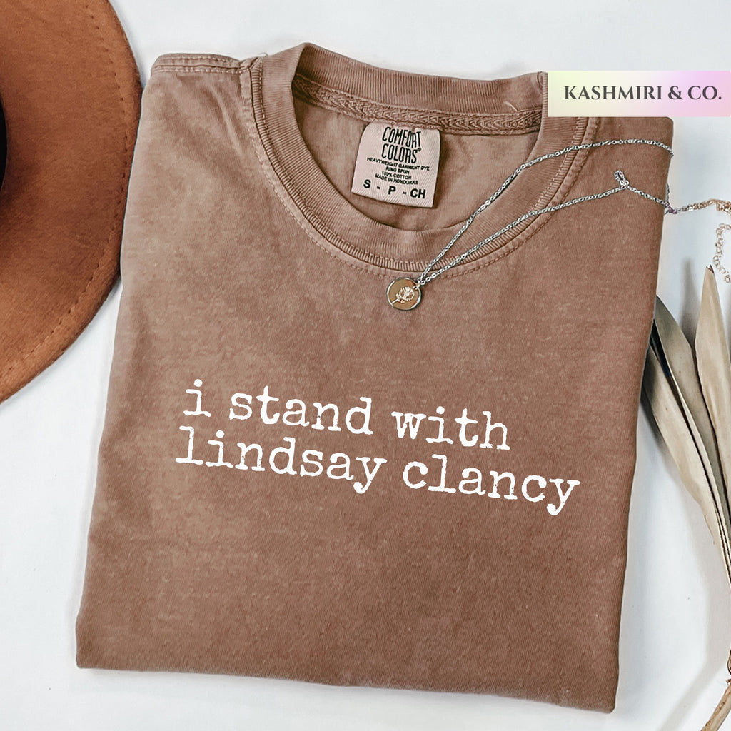 A folded brown t-shirt with white printed text reading 'i stand with lindsay clancy' on a white background, styled with a necklace, a brown hat, and a book. A brand tag for 'Comfort Colors' is visible inside the collar, and a 'KASHMIRI & CO.