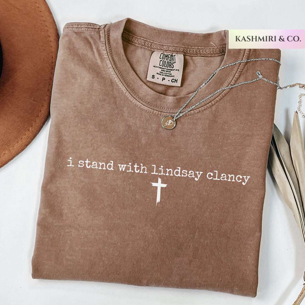 A folded brown t-shirt with white text reading 'i stand with lindsay clancy' and a white cross, styled with a necklace and a partial hat on a white background. A brand tag for 'Comfort Colors' is visible inside the collar, and a 'KASHMIRI & CO.