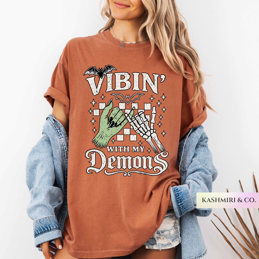 A woman wearing an oversized rust-brown graphic T-shirt with rolled sleeves, layered with a light blue denim jacket. The shirt features a gothic-style graphic with the text 'VIBIN' WITH MY DEMONS' and illustrations of a bat, a green hand, and a skeleton hand.