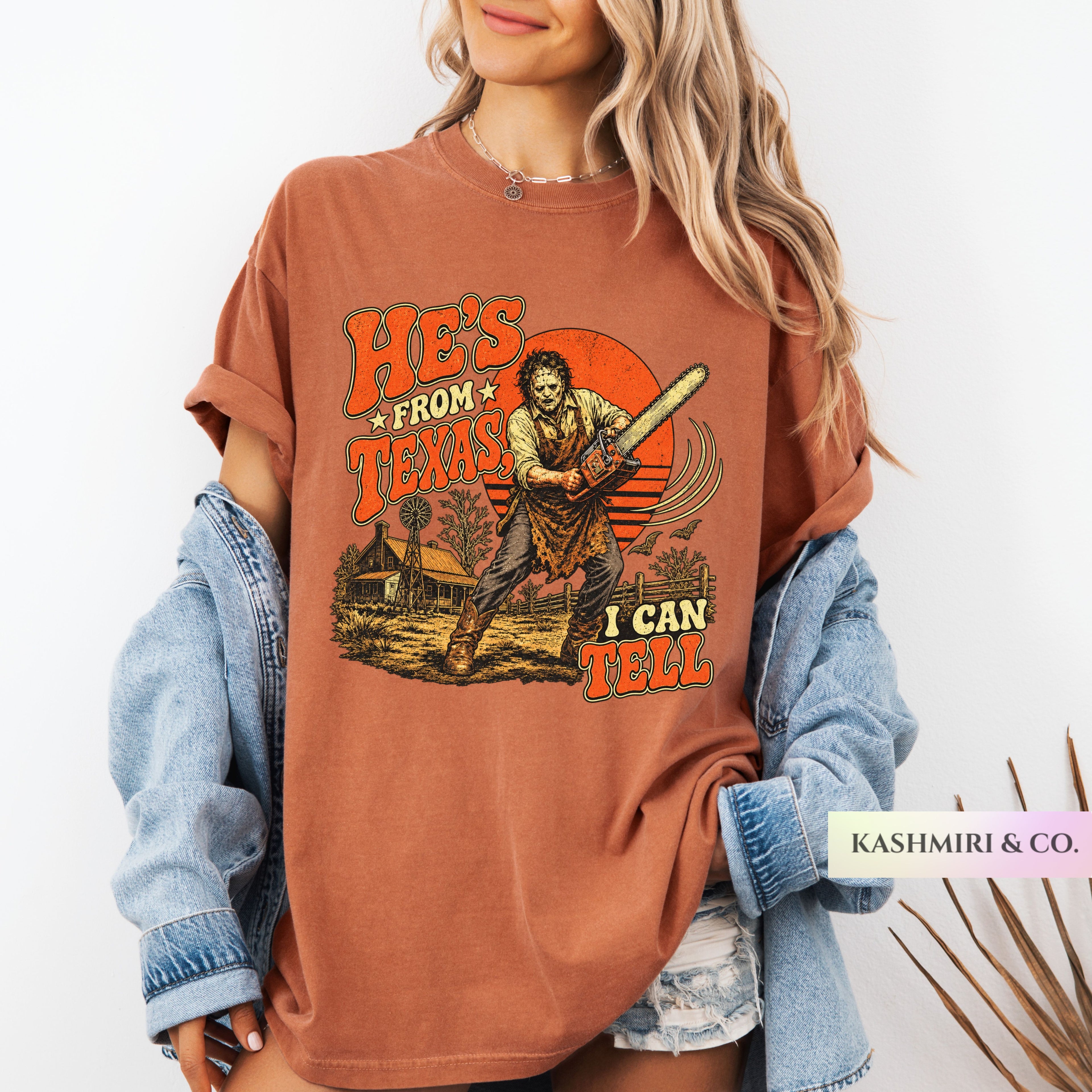 A woman wearing an oversized rust-brown graphic T-shirt with a horror-themed print reading 'HE'S FROM TEXAS I CAN'T TELL' and showing a character holding a chainsaw bat. She has a light blue denim jacket off her shoulders and is standing against a plain white background.