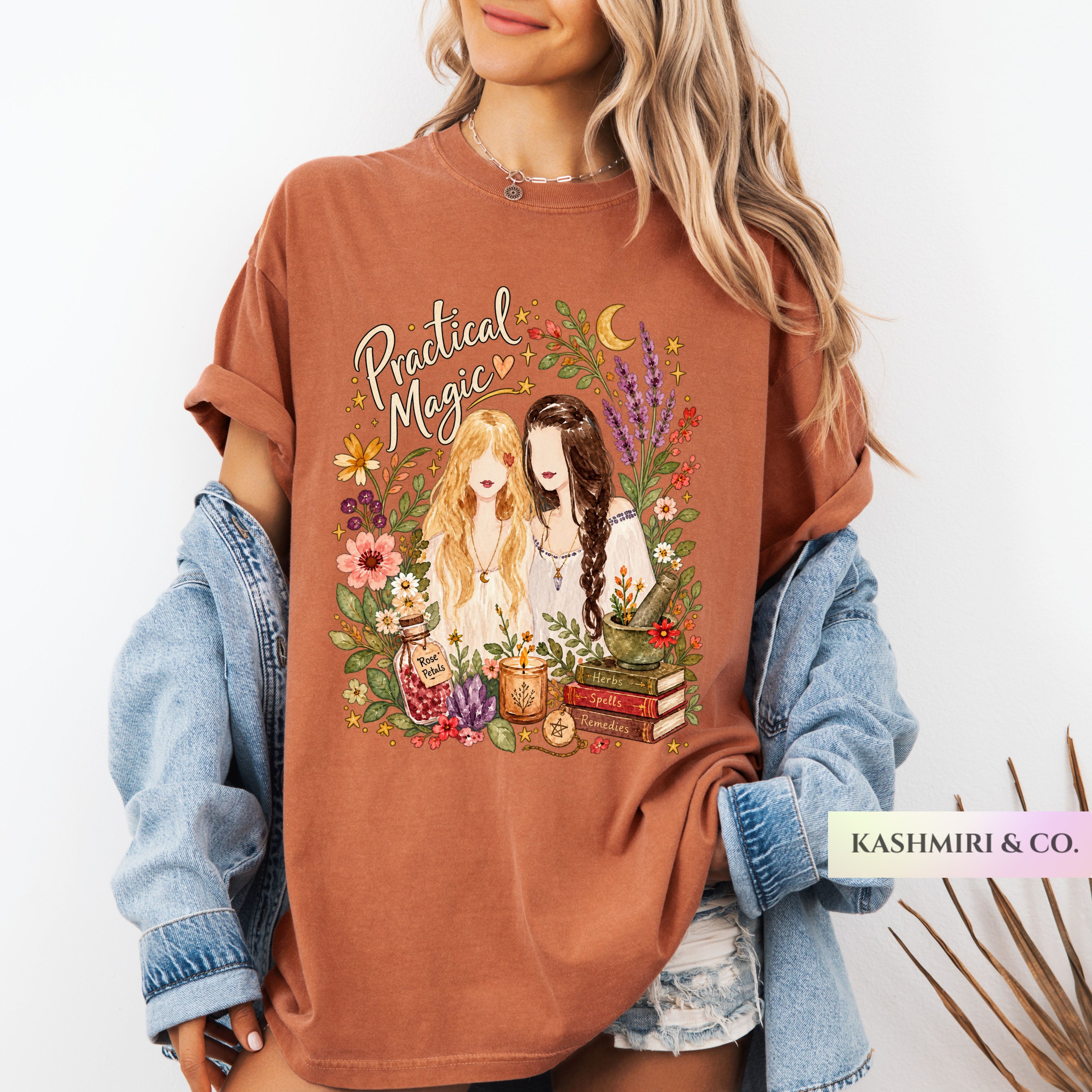 A woman wearing an oversized rust-brown graphic T-shirt with the words 'Practical Magic' and an illustrated floral scene, styled with a light blue denim jacket and denim shorts against a white background.