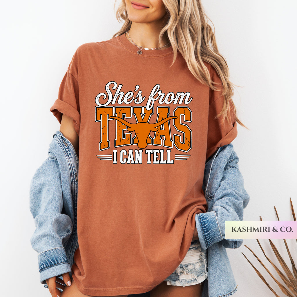 A woman wearing an oversized rust-brown graphic T-shirt with rolled sleeves and a light blue denim jacket draped off her shoulders. The shirt features the text 'She's from TEXAS I CAN TELL' with a longhorn silhouette.
