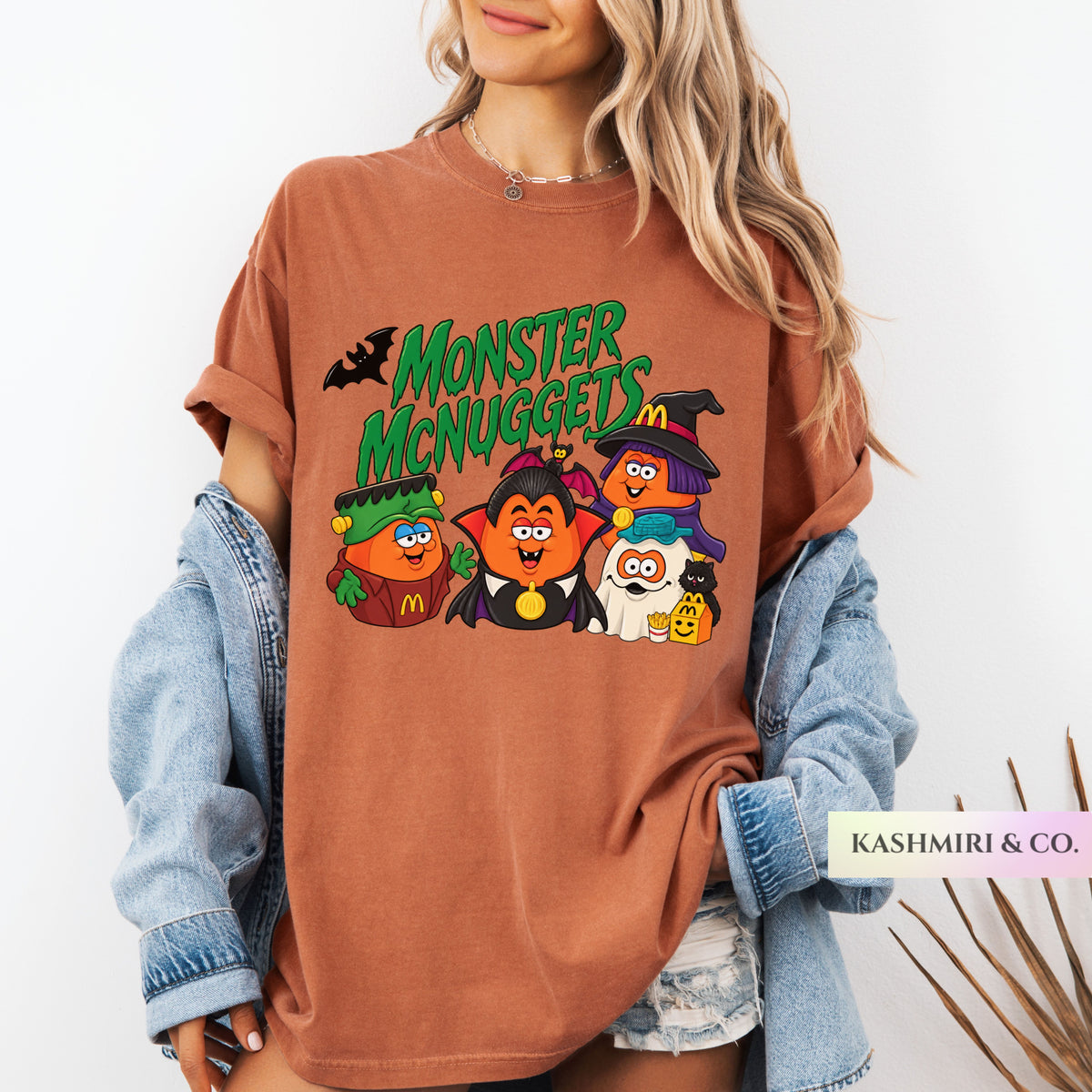 A woman wearing an oversized brown graphic T-shirt with a colorful 'Monster McNuggets' Halloween print, styled with a light blue denim jacket and denim shorts against a white background.