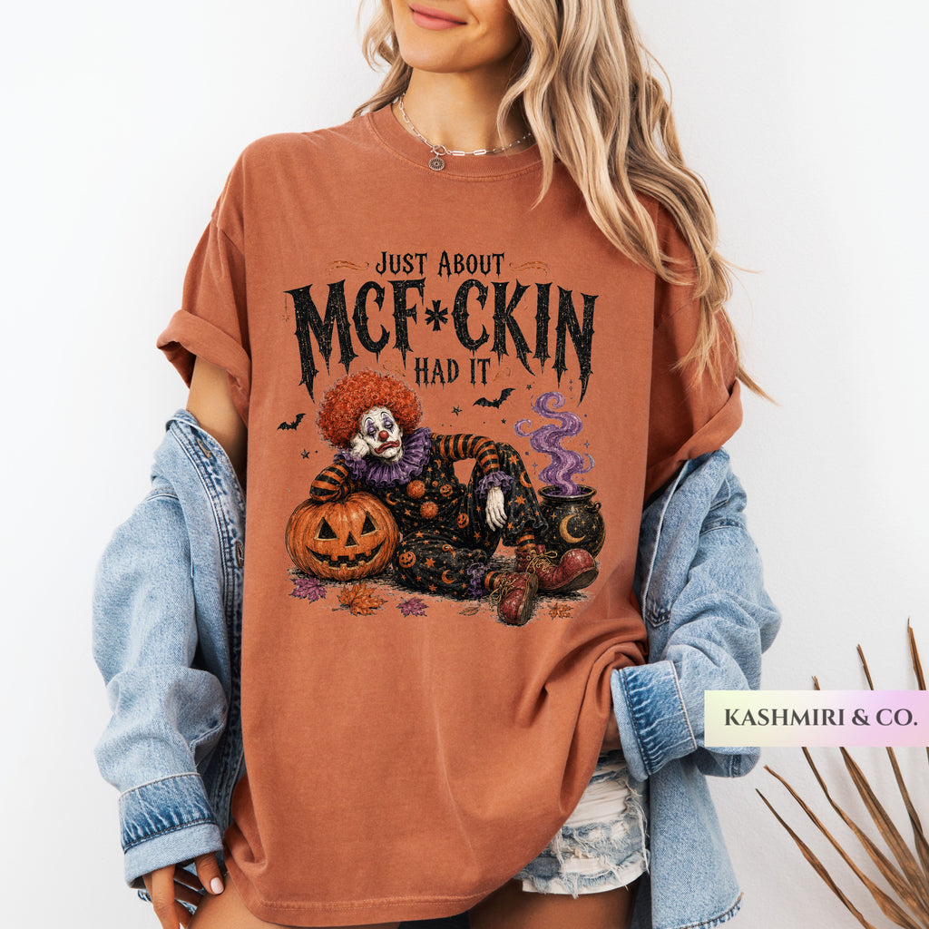 A woman wearing an oversized rust-orange graphic T-shirt with a Halloween clown illustration and the text 'JUST ABOUT MCF*CKIN HAD IT,' styled with a light blue denim jacket and denim shorts against a plain white background.