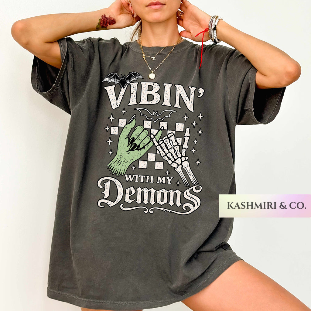 A woman wearing an oversized dark gray graphic t-shirt with a Halloween-themed print that reads 'VIBIN' WITH MY Demons' featuring a green hand, skeleton hand, and bats. A 'KASHMIRI & CO.