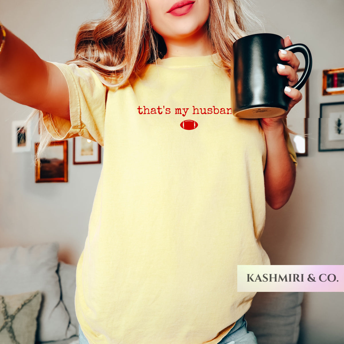 A woman wearing a pale yellow graphic t-shirt with the red text 'that's my husband' taylor swift travis kelce and a small football icon, holding a black ceramic mug in a cozy indoor setting. The brand name 'KASHMIRI & CO.