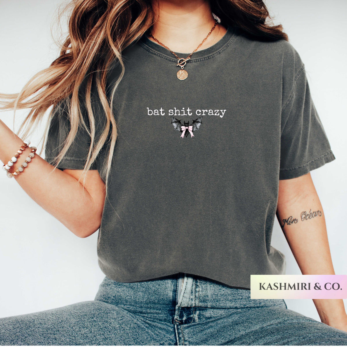 A woman wearing a dark gray washed graphic t-shirt with the text 'bat shit crazy" bat and a bow illustration. The brand name 'KASHMIRI & CO.