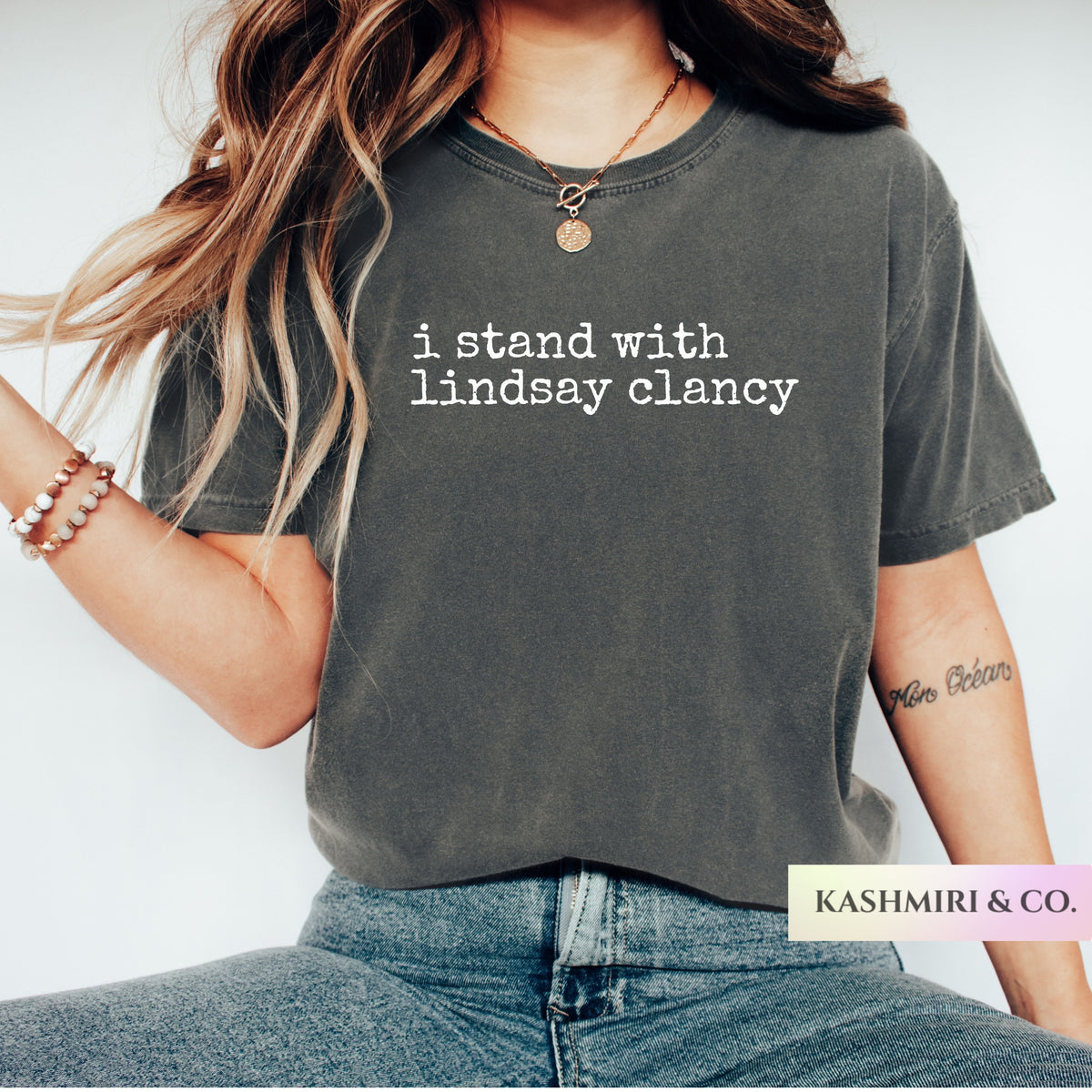 A woman wearing a dark charcoal gray graphic t-shirt with the white text 'i stand with lindsay clancy' printed on the chest. The brand label 'KASHMIRI & CO.