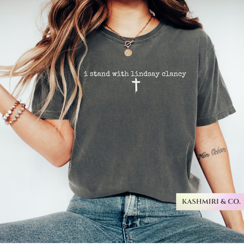 A woman wearing a charcoal gray graphic t-shirt with the white printed text 'i stand with lindsey clancy' and a white cross, paired with blue jeans. The brand label 'KASHMIRI & CO.