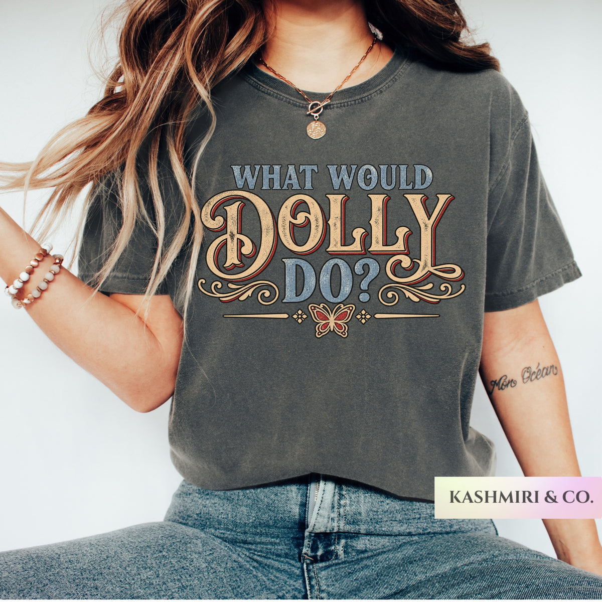 A woman wearing a dark gray graphic t-shirt with the text 'WHAT WOULD DOLLY DO?' in blue and gold lettering, paired with blue jeans. The brand name 'KASHMIRI & CO.