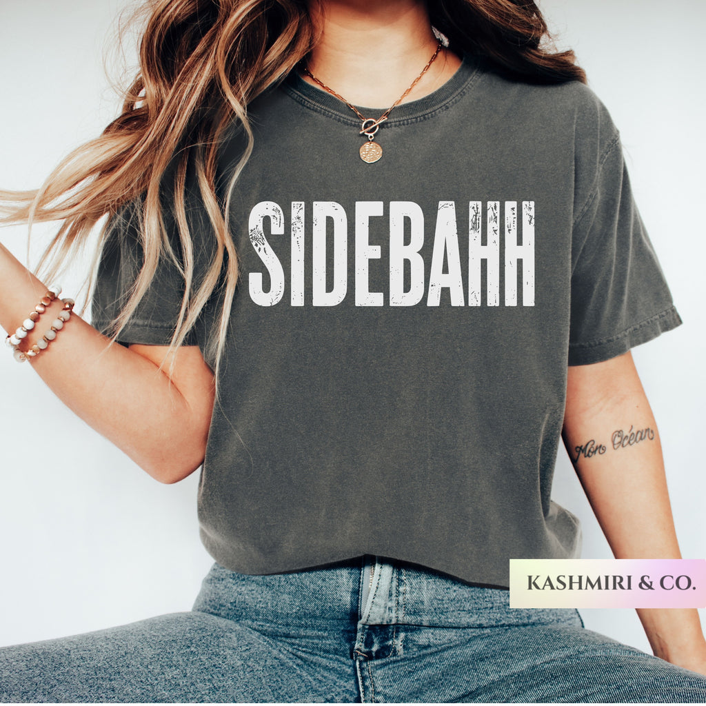 A woman wearing a dark gray washed graphic t-shirt with the word 'SIDEBAHH' printed in distressed white capital letters across the chest. She is also wearing blue jeans, a gold necklace, and bracelets.