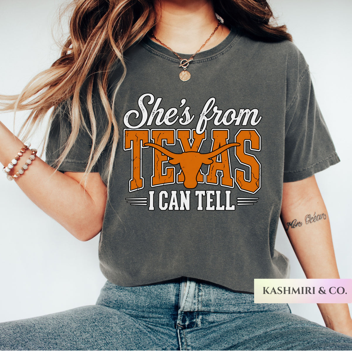 A woman wearing a dark gray graphic t-shirt with the text 'She's from TEXAS I CAN TELL' and a longhorn steer graphic. The brand name 'KASHMIRI & CO.