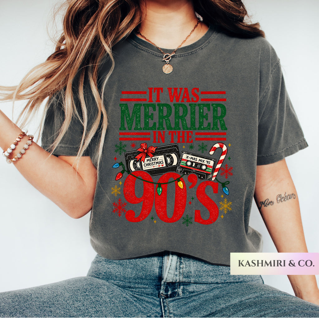 A woman wearing a dark gray graphic t-shirt with a '90s Christmas' design, paired with blue jeans. The graphic features retro cassette tapes, candy canes, and colorful Christmas lights.