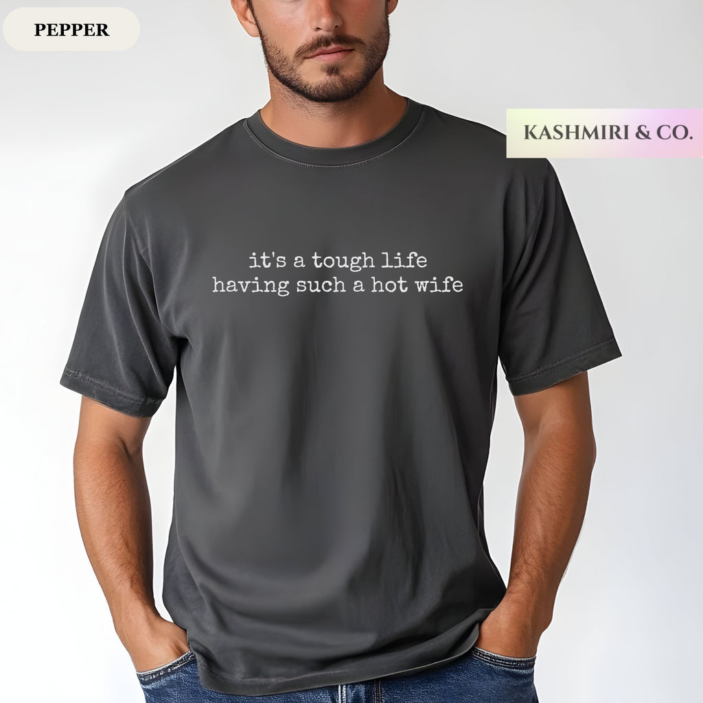 A man wearing a dark gray 'pepper' t-shirt with white printed text that reads 'it's a tough life having such a hot wife'. The brand logo 'KASHMIRI & CO.