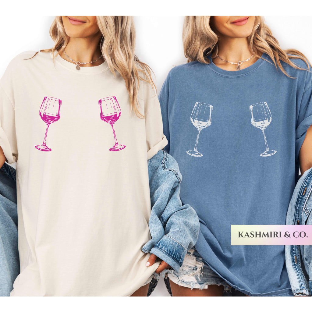Two women modeling oversized graphic T-shirts side by side. The left model wears an off-white/cream shirt with two pink wine glass illustrations, and the right model wears a dusty blue shirt with two white wine glass illustrations.