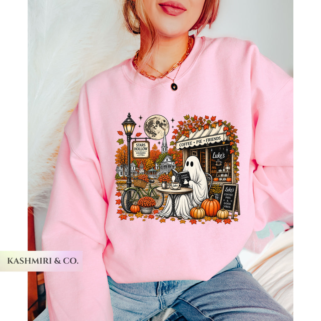 A woman wearing a light heather gray sweatshirt with a detailed autumn-themed graphic print featuring a ghost, pumpkins, a street lamp, and a moon. Stars Hallow Luke's Diner